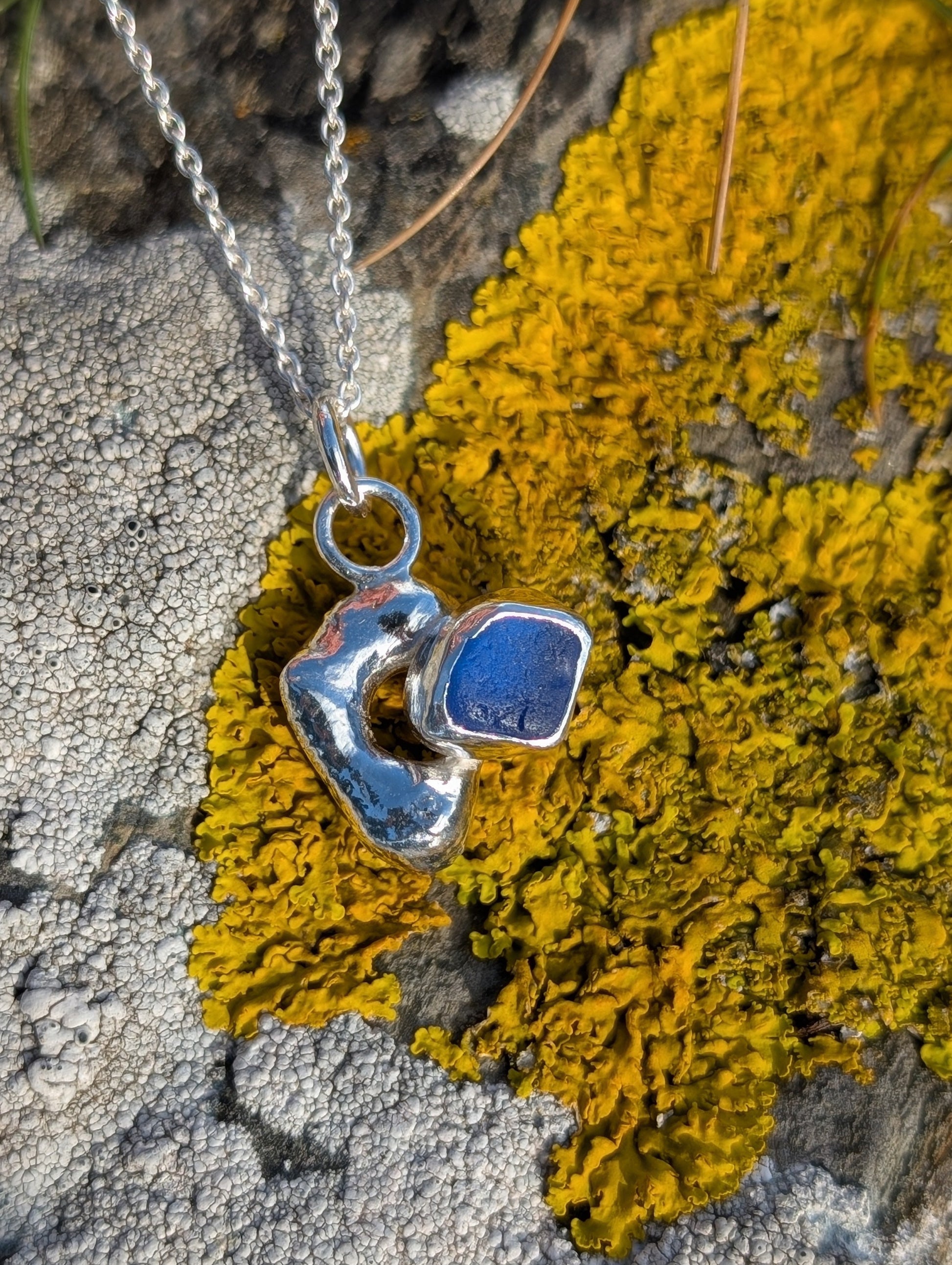 Silver pendant with blue stone on a chain, lying on yellow lichen.